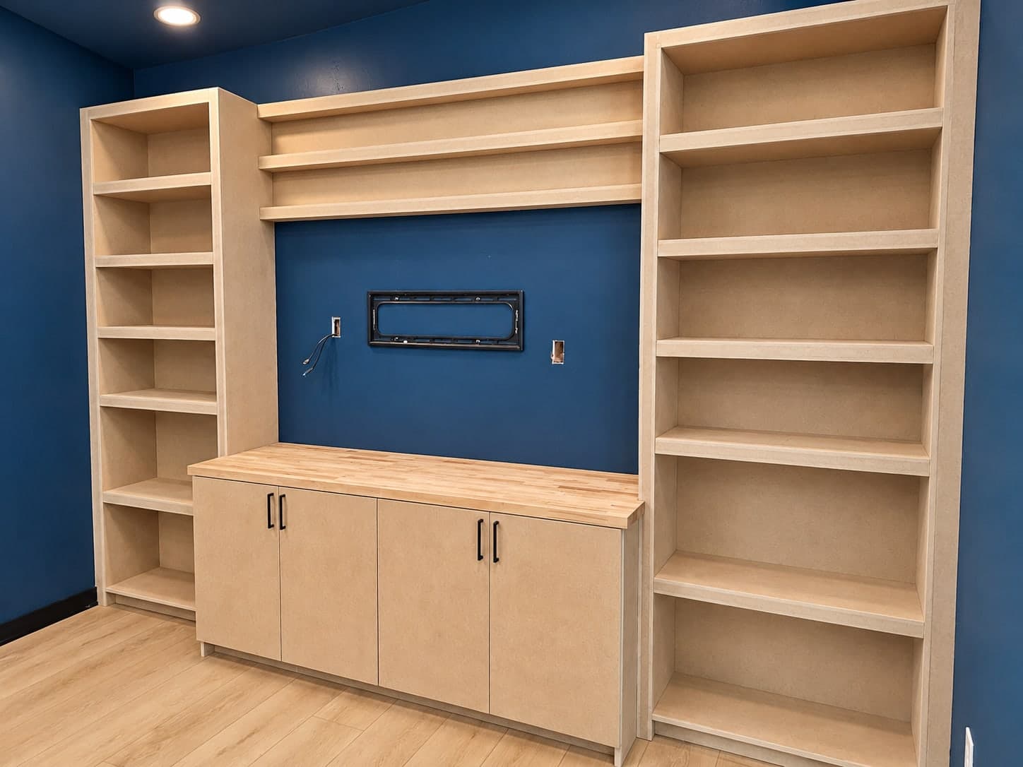 Built-in bookcases in unfinished MDF on both sides of a navy accent wall with a TV mount, with open shelves above and a butcher block topped cabinet below