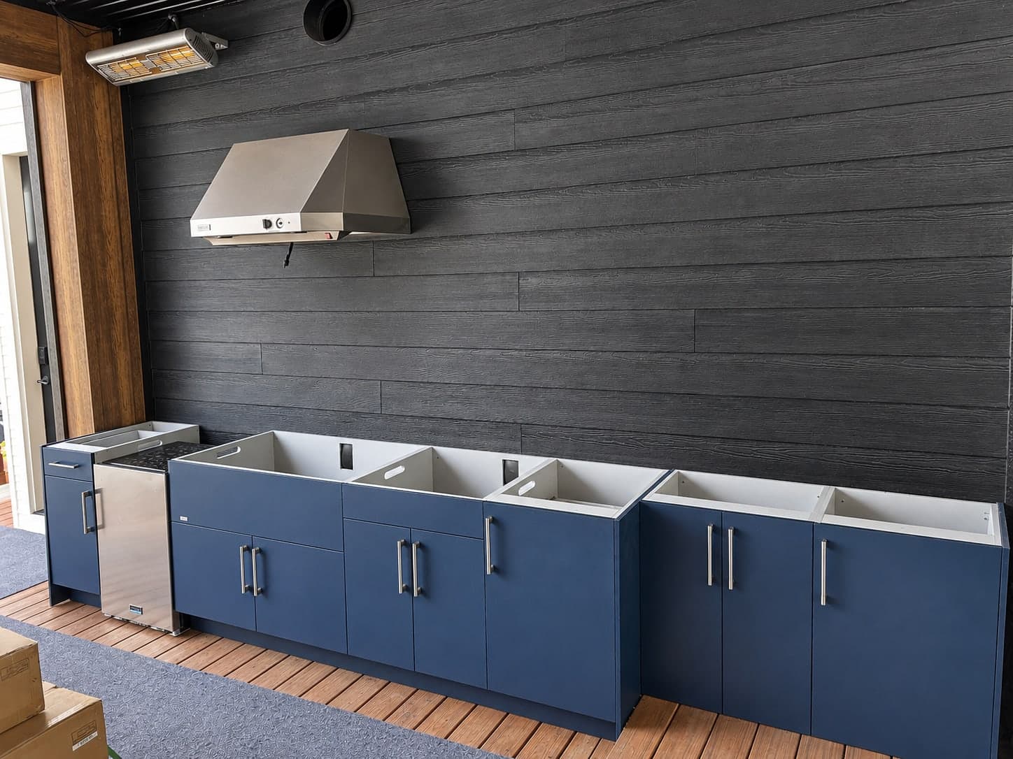 Navy outdoor kitchen cabinets with steel bar pulls and a stainless under-counter fridge below a stainless range hood, on a covered deck with dark wood-grain siding and a wall-mounted heater