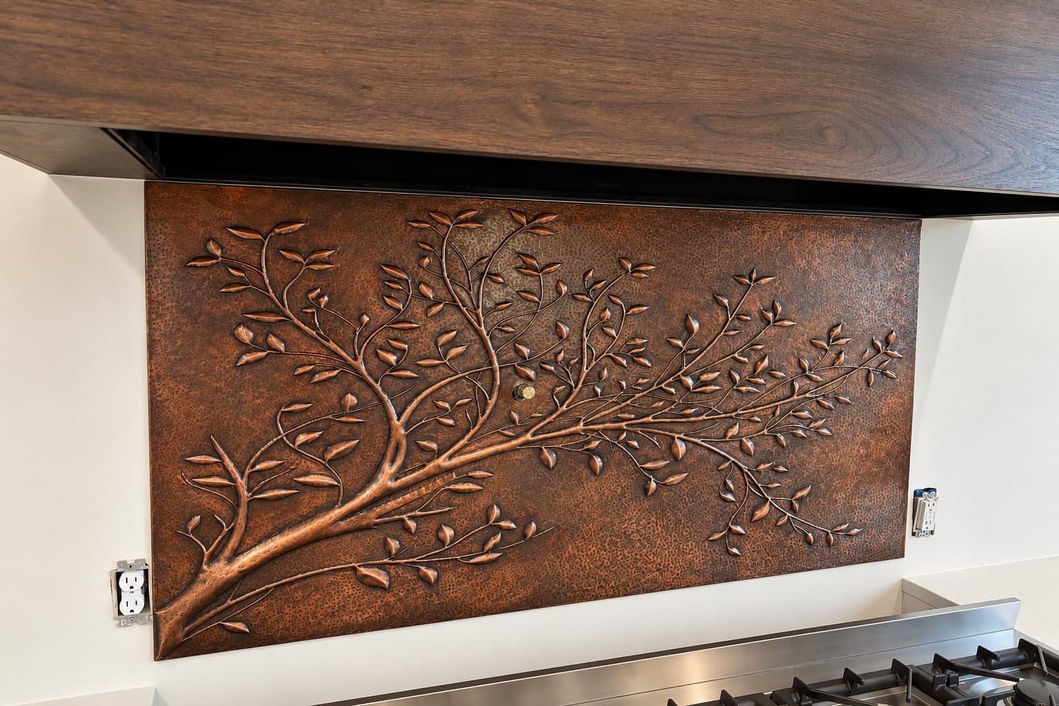 Hammered copper backsplash panel with a tree branch in relief, between a wood range hood and a stainless range