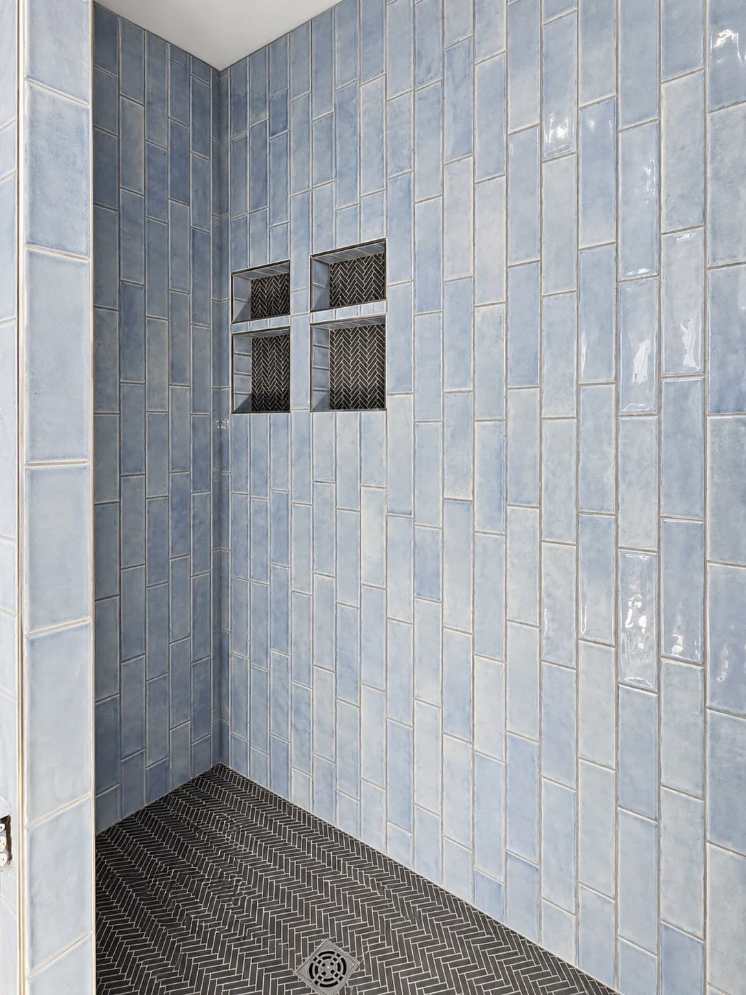 Side view of the blue tile shower, with recessed niches backed in black herringbone tile above a black herringbone floor