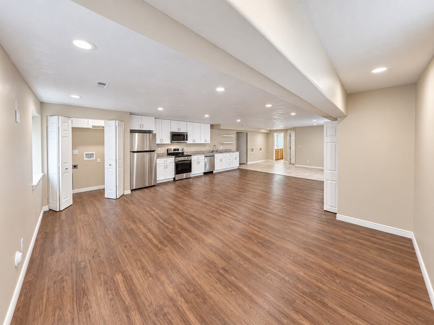 Open finished basement with wood-look plank flooring, a laundry closet with bifold doors, and a kitchenette of white cabinets with a stainless fridge, range, microwave and dishwasher