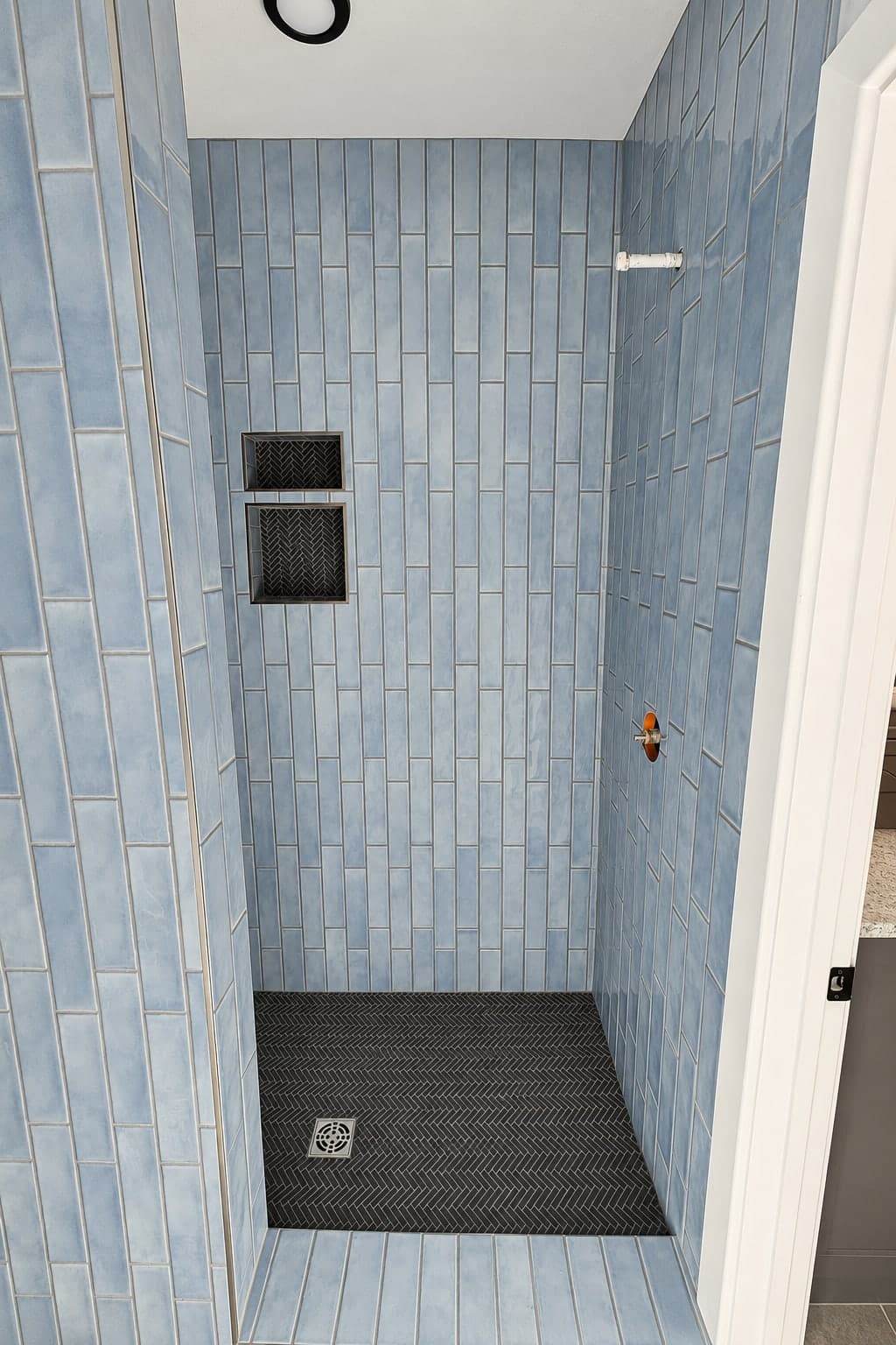 Walk-in shower in vertical blue tile with recessed niches and a black herringbone floor