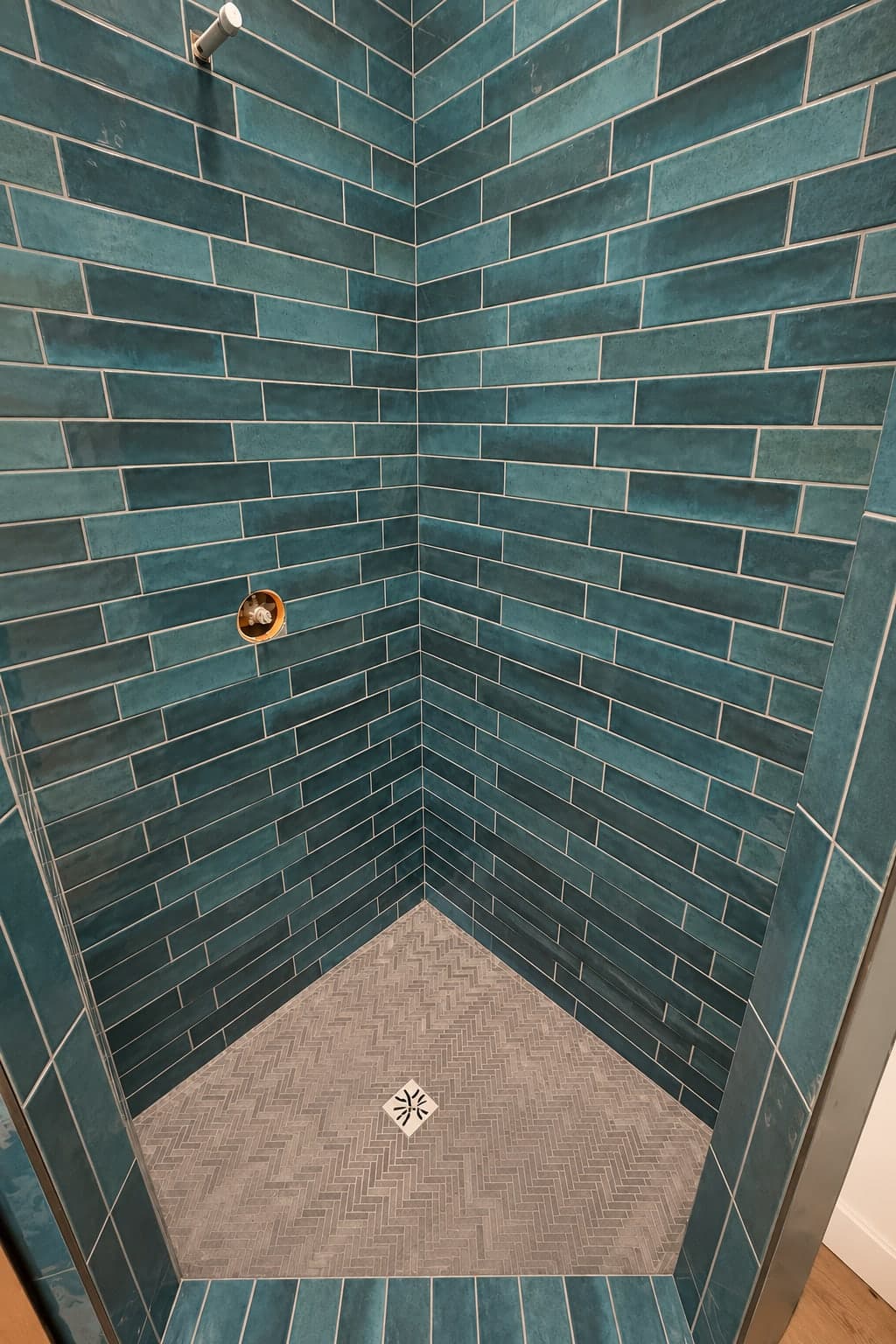 Corner shower in long teal glazed tile with a gray herringbone floor and a square decorative drain