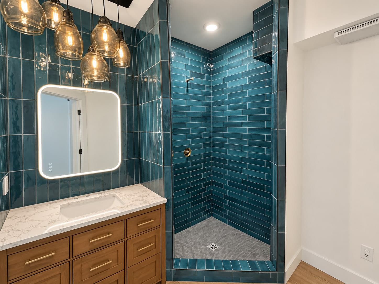 Bathroom with a wood vanity, white counter and backlit mirror under amber glass pendant lights against teal tile, beside the teal tile corner shower