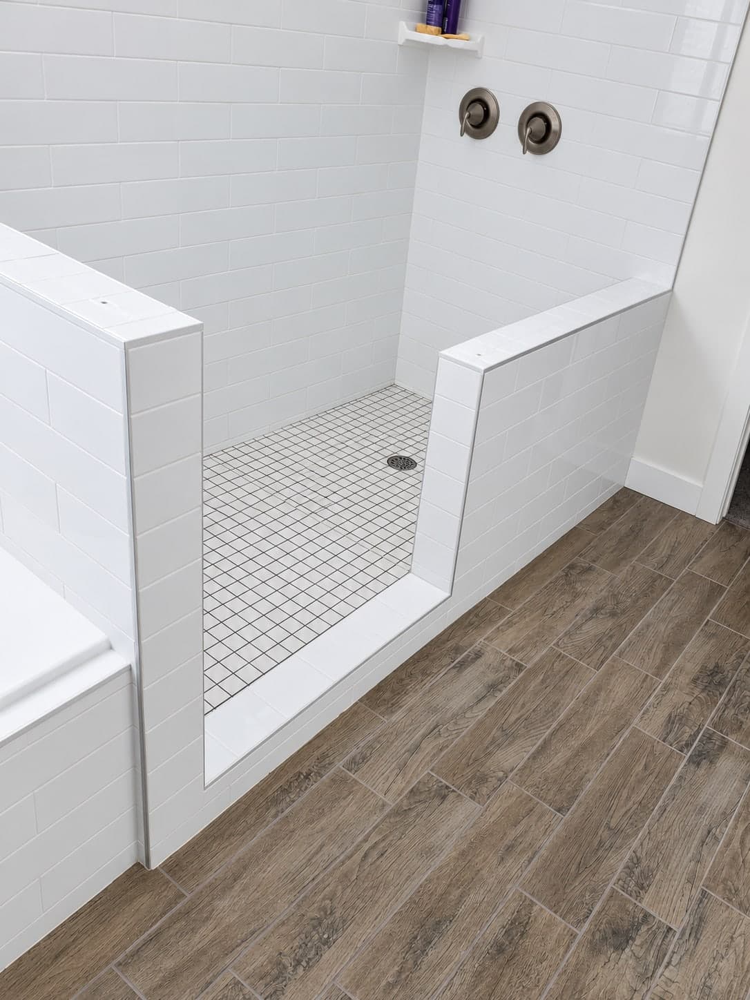 Walk-in shower with white subway tile half walls and a white square mosaic floor with a center drain, next to a tiled tub, above a wood-look plank tile floor