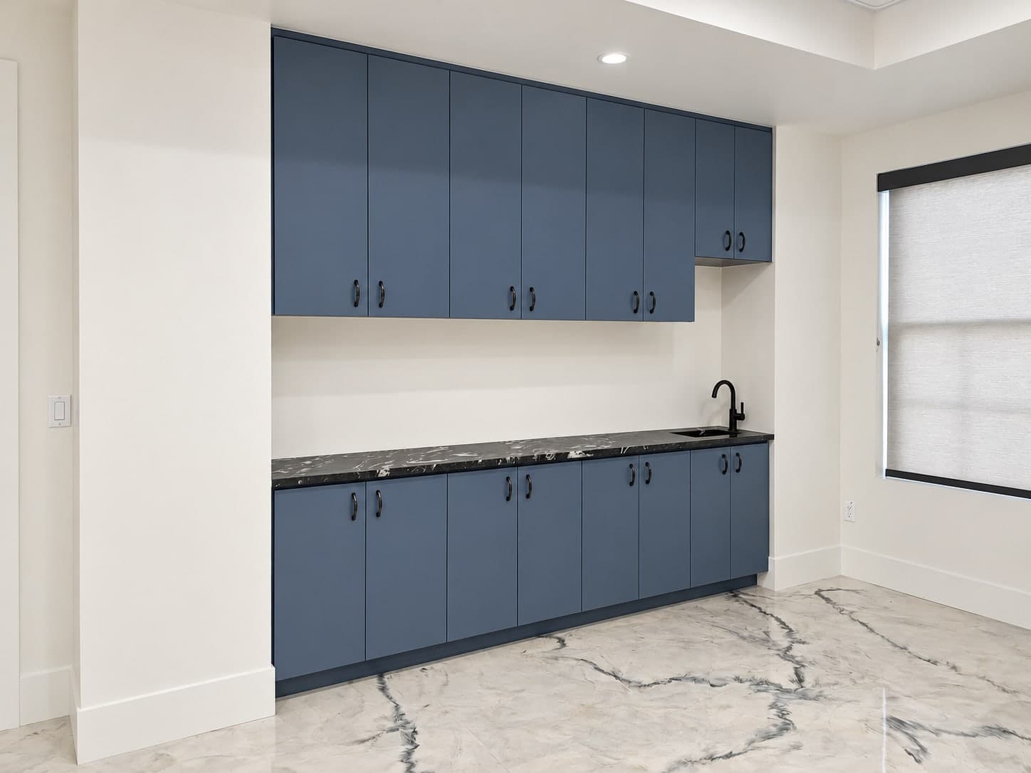 Blue upper and lower cabinets around a dark stone counter with a sink and a black faucet, beside a window with a roller shade