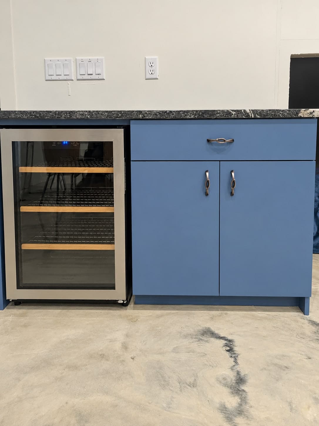 Stainless wine fridge beside a blue base cabinet with a drawer and two doors, under a dark stone counter
