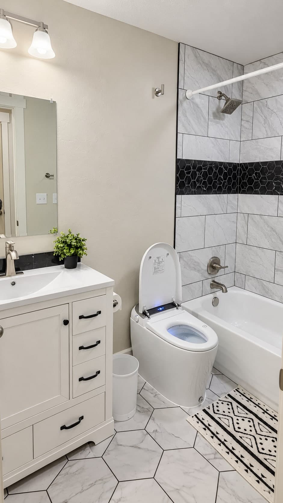 Bathroom with a white vanity with black pulls and a black stone backsplash, a smart toilet with its lid open, and the gray tile tub and shower with its black hexagon band, over a white hexagon tile floor