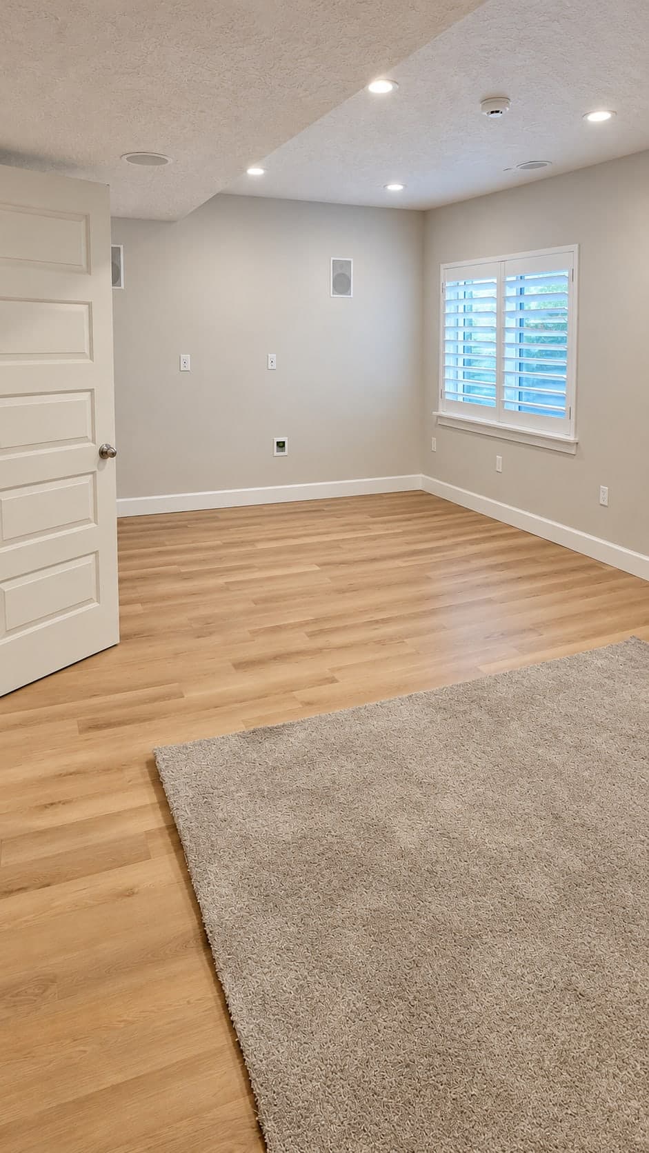 The same room toward its shuttered window, with plank flooring, a rug and in-wall speakers