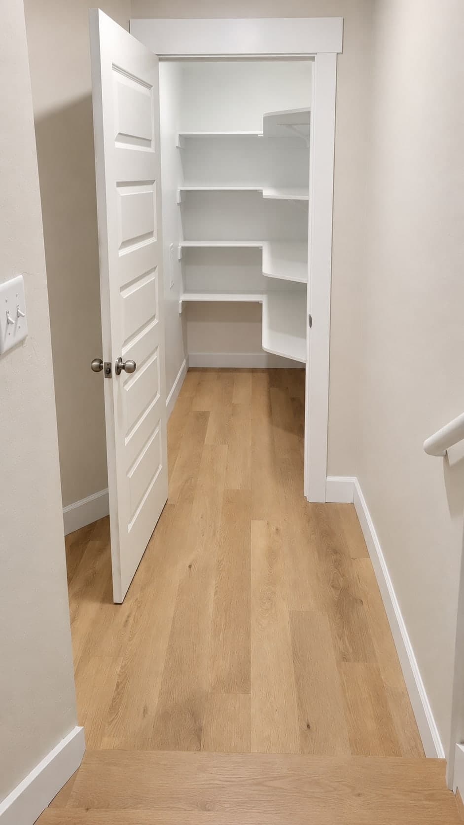 Storage room with white wraparound shelves behind a paneled door, off a hall with plank flooring beside the stairs