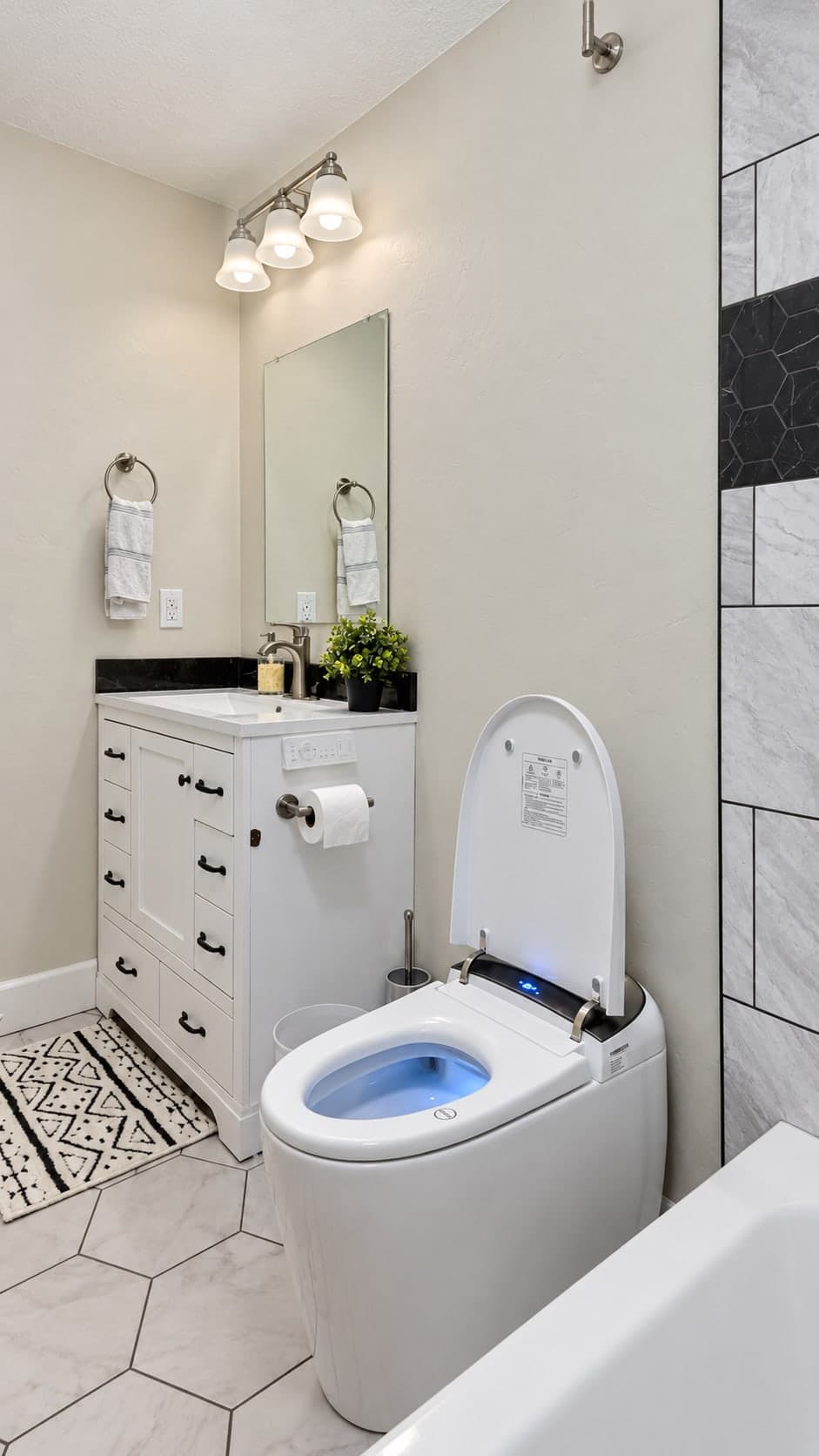 White vanity with black pulls and a black stone backsplash under a mirror and a three-light fixture, beside a smart toilet with its lid open, on a white hexagon tile floor